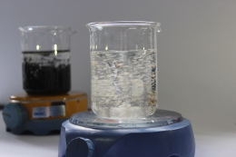 Ein Glasbehälter mit klarem Wasser auf einem Hotplate, im Hintergrund ein weiterer Glasbehälter mit schwarzer Flüssigkeit.